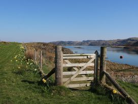 A wooden gate on a grassy path by a wire fence with yellow flowers beside water and hills at Insh Cullipool Isle Of Luing