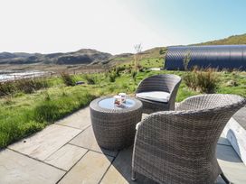 Two wicker chairs and a wicker table with two mugs on a stone patio overlooking grassy hills and a corrugated metal structure at Sula Cullipool Isle Of Luing