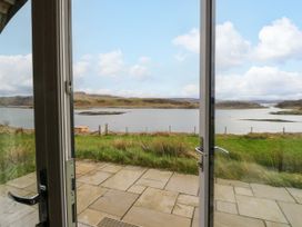 View through open glass doors onto a stone patio and grassy area overlooking a loch with hills in the distance at Losal Cullipool Isle Of Luing
