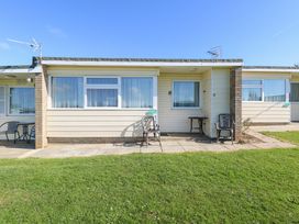 A single story building with windows chairs and tables on a stone patio in front of a lawn at Chalet 37 in Hemsby
