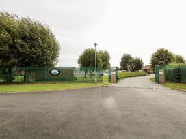 A gated entrance with green metal fencing and a paved road leading to buildings and trees at Chalet 37 in Hemsby