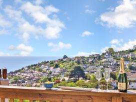 A wooden railing with a bowl of snacks a plate two champagne glasses and a bottle of champagne with a view of houses on a hillside and the sea at Seascape Cottage in Beer