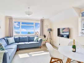 A living room with a blue corner sofa a round white table with a bottle and glasses a wall mounted tv and a window with a sea view at Beer View Cottage in Beer