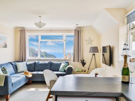 A living room with a blue corner sofa a window showing a sea view a floor lamp a wall-mounted TV and a kitchen counter with a bottle of champagne at Beer View Cottage in Beer