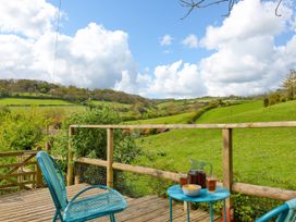A wooden deck with blue chairs a table with drinks and a bowl overlooking green hills and cloudy sky at Beer View Cottage in Beer