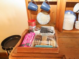 A wooden tray with biscuits coffee packets and a jar on a wooden surface with blue mugs and plates in a wooden cupboard at Pagecroft Cottage in Haydon Bridge