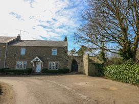 A stone cottage with a white door and windows a gravel driveway and a gate next to a large tree at Pagecroft Cottage in Haydon Bridge