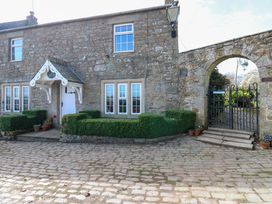 A stone cottage with windows and a white door next to a gated stone archway and cobblestone driveway at Pagecroft Cottage in Haydon Bridge