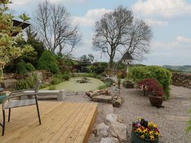 A garden with a pond surrounded by plants and trees and a patio with chairs at Pagecroft Cottage in Haydon Bridge