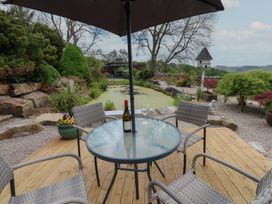 An outdoor seating area with a glass table, four chairs, an umbrella, a pond, and garden plants at Pagecroft Cottage in Haydon Bridge