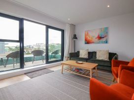 A living room with green sofa, orange chairs, wooden coffee table, floor lamp, abstract painting, and balcony with chairs at Ebbtide in Newquay