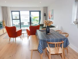 A dining area with wooden chairs and table with patterned tablecloth next to a living room with red armchairs and sliding glass doors at Ebbtide in Newquay