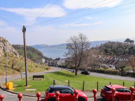 A coastal landscape with a road, parked cars, a grassy area with a bench and a tree, houses, hills, and water in the background at 4 Bay View Terrace in Llandudno