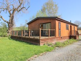 A wooden lodge with a deck and railing surrounded by grass and trees at Chestnut Lodge in Clunton near Clun