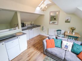 A small kitchen area with white cabinets and appliances next to a living area with a grey sofa and colorful cushions at Cuddfan at Swallow Cottage in Rhydtalog near Coedpoeth