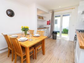 A kitchen dining area with a wooden dining table set for four, a vase of yellow flowers, wooden chairs, a wall clock, kitchen cabinets, a trash bin, and glass double doors at Cape 12 Near Yarmouth
