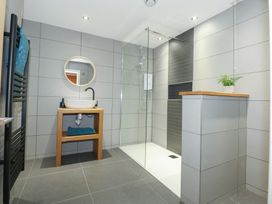 A bathroom with a wooden sink stand and round mirror next to a walk in shower and a tiled half wall with a potted plant at Criw in Bryncroes near Sarn Meyllteyrn