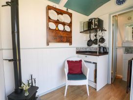 A small kitchen area with a white chair with red and blue cushions a stove a wall-mounted wooden cupboard with plates and a microwave on a shelf at Songbirds in St Michaels Kent