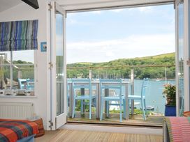 A room with an open glass door leading to a balcony with blue chairs and a table overlooking a river and green hills at 5 Esplanade in Fowey