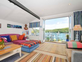A living room with a purple sofa, colorful cushions, striped rugs, a coffee table, and an open door leading to a balcony with blue chairs overlooking water at 5 Esplanade in Fowey