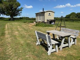 A wooden table with benches on grass near a small shed with a tree and blue sky at Shepherd Hut 1 in Northchapel