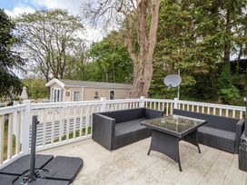 An outdoor patio with black wicker furniture and a glass table near a beige cabin surrounded by trees at 155 Woodland Hall Lyons in Ruthin