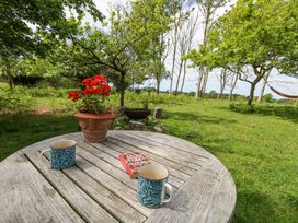 A wooden outdoor table with two mugs a book and a flowerpot in a garden with trees and a hammock at Fallow in Warmwell