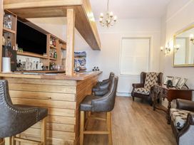 A wooden home bar with stools and liquor bottles and a sitting area with armchairs and a table with picture frames at The Burleigh in Hunstanton