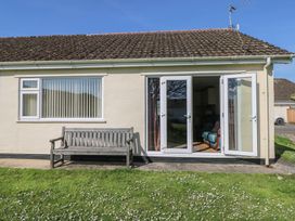 The exterior of a single-story house with a wooden bench on a concrete path and open double glass doors showing a blue armchair inside at Kites - 52 Gower Holiday Village in Scurlage near Port Eynon and Rhossili