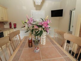 A kitchen dining area with a wooden table and chairs a vase of pink flowers bottles and coasters on the table a wall-mounted television and a mirror on the wall at Killelton Cottage in Camp County Kerry