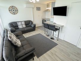 A living room with two black sofas a wall clock and a kitchen area with bar stools and a wall-mounted tv at 99 Cherry Park in Chapel St Leonards