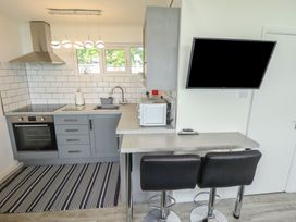 A kitchen with gray cabinets a stovetop oven a sink a microwave and two black bar stools at a counter with a wall-mounted TV at 99 Cherry Park in Chapel St Leonards