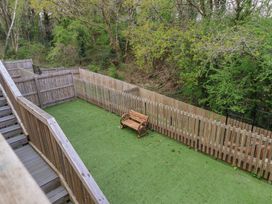 A fenced garden area with artificial grass and a wooden bench surrounded by trees at Peninsula Cottages - The Tors in Tavistock