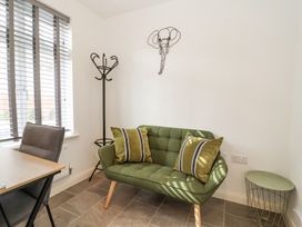 A small room with a green sofa and two pillows a coat rack a desk and chair by a window with blinds and a small round table Peninsula Cottages - The Tors Tavistock