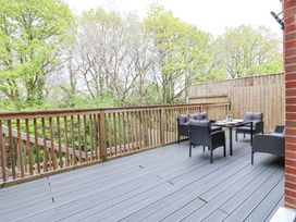 An outdoor wooden deck with black cushioned chairs around a glass table surrounded by wooden railings and trees in the background at Peninsula Cottages - The Tors in Tavistock