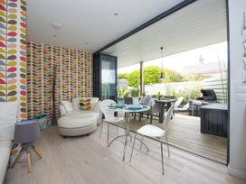 A small dining area with a round table and two white chairs next to a seating area with a round couch and patterned wallpaper opening to an outdoor patio with a dining table and barbecue at Two Bare Feet Rose near Perranporth