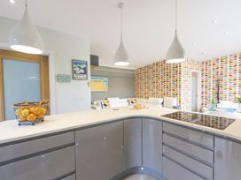 A kitchen with a curved island counter and two hanging lights a living area with white sofas and patterned wallpaper at Two Bare Feet Rose near Perranporth