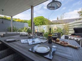 An outdoor covered dining area with a table set with plates glasses a bottle of wine and snacks next to a barbecue grill and surfboards at Two Bare Feet Rose near Perranporth