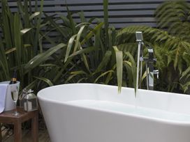 An outdoor bathtub with water flowing from a faucet surrounded by tall green plants and a wooden table holding a wine glass a bottle in an ice bucket and a lantern at Two Bare Feet Rose near Perranporth