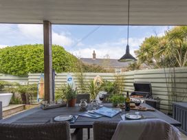 An outdoor dining area with a table set with wine glasses and a bottle next to a barbecue grill and surfboards near the fence at Two Bare Feet in Rose near Perranporth