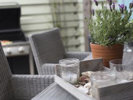 A patio table with a wooden tray holding glass jars and seashells next to a potted lavender plant with a barbecue and wicker chairs in the background at Two Bare Feet Rose near Perranporth