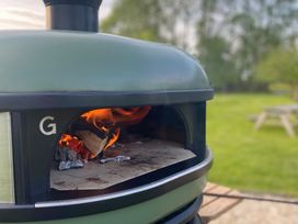 An outdoor wood fire oven with flames inside and a green lawn with a picnic table in the background at Pippin lodge in Coxbridge near Glastonbury