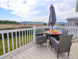 An outdoor deck with a round table set for dining four gray wicker chairs a closed umbrella and a rural landscape view at Lawi39 in Towyn