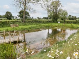 A pond with a wooden deck and bench surrounded by trees and flowers at Little Acorn Cottage in Munsley near Ledbury