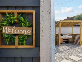 A welcome sign with green leaves on a dark door next to a patio area with a grill under a wooden pergola at Fishermans Cottage in Prestatyn