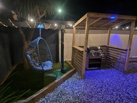 An outdoor area at night with a hanging cushioned chair, string lights, a wooden pergola with a barbecue grill, and gravel ground