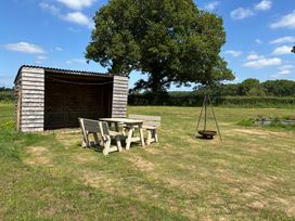 A picnic table with benches on a grassy lawn near a wooden shelter and a fire pit in a field at Shepherds Hut 2 in Northchapel