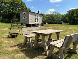 A picnic table with benches and a fire pit in front of a shepherds hut in a grassy area with trees at Shepherds Hut 2 in Northchapel