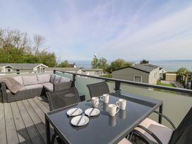 An outdoor balcony with a glass table set with cups and plates and a corner sofa overlooking cabins and the sea at Heulwen in New Quay