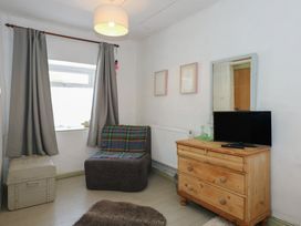 A small room with a window covered by gray curtains a plaid-covered chair a wooden chest of drawers with a TV on it and a mirror on the wall at Cobweb Cottage in Waunfawr